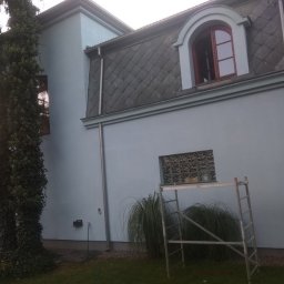 Arkadiusz Matuszak - Niebieski budynek z szarym dachem, widoczna aluminiowa drabina oparta o ścianę pod oknem z luksferami, zarośnięty drzewem fragment fasady.