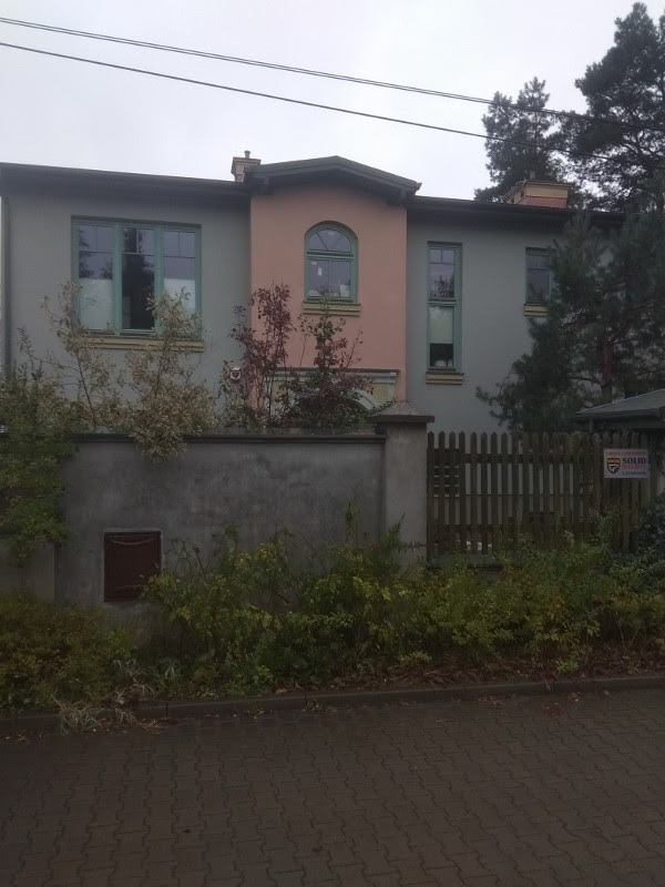 Dwupiętrowy dom z szarą elewacją i różową wstawką nad oknem, ogrodzony betonowym murem z drewnianym płotem po prawej stronie, zarośnięty krzewami.