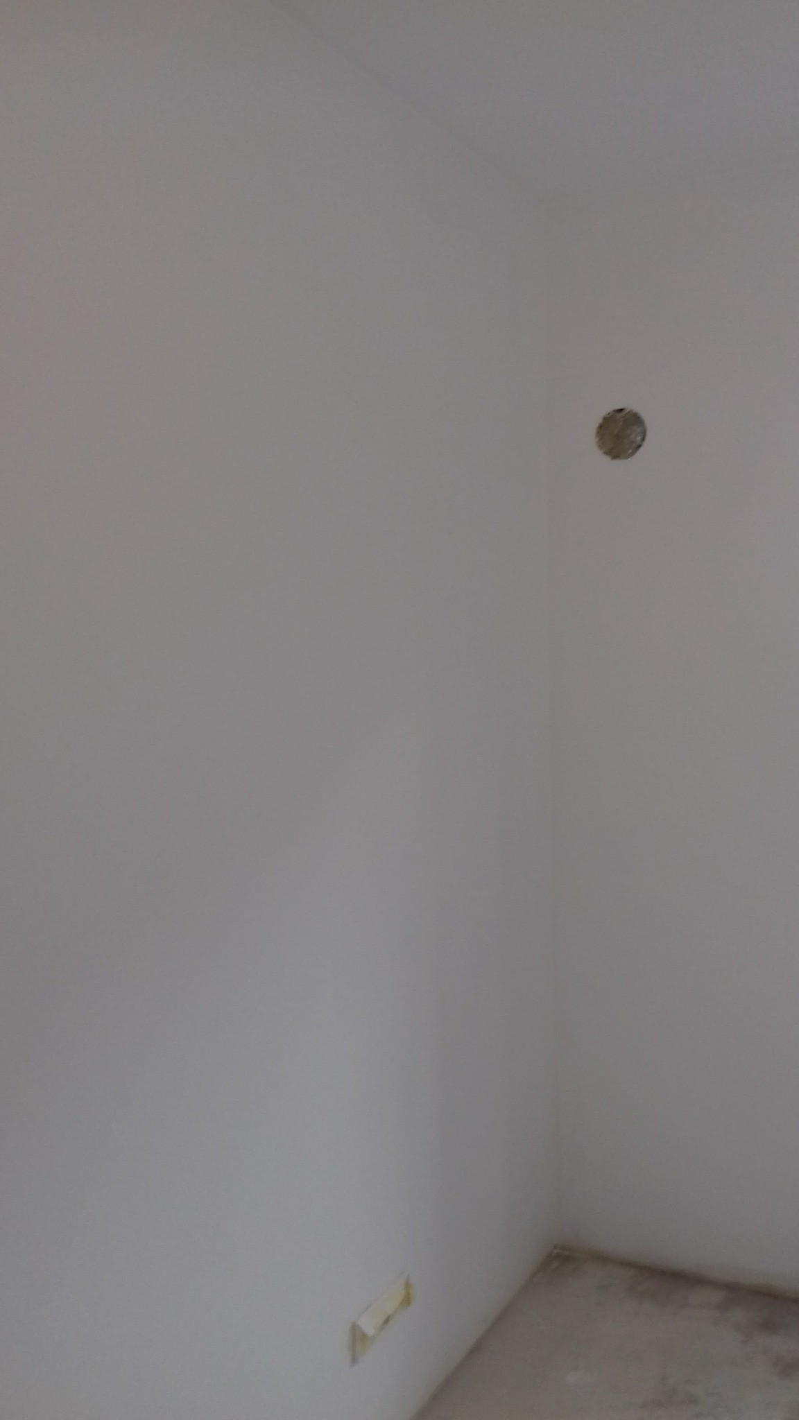 Wnętrze w trakcie prac przygotowawczych: gładka, biała ściana z widocznym otworem na instalację elektryczną w górnej części i zabezpieczonym gniazdkiem w dolnej części, surowa podłoga w rogu...