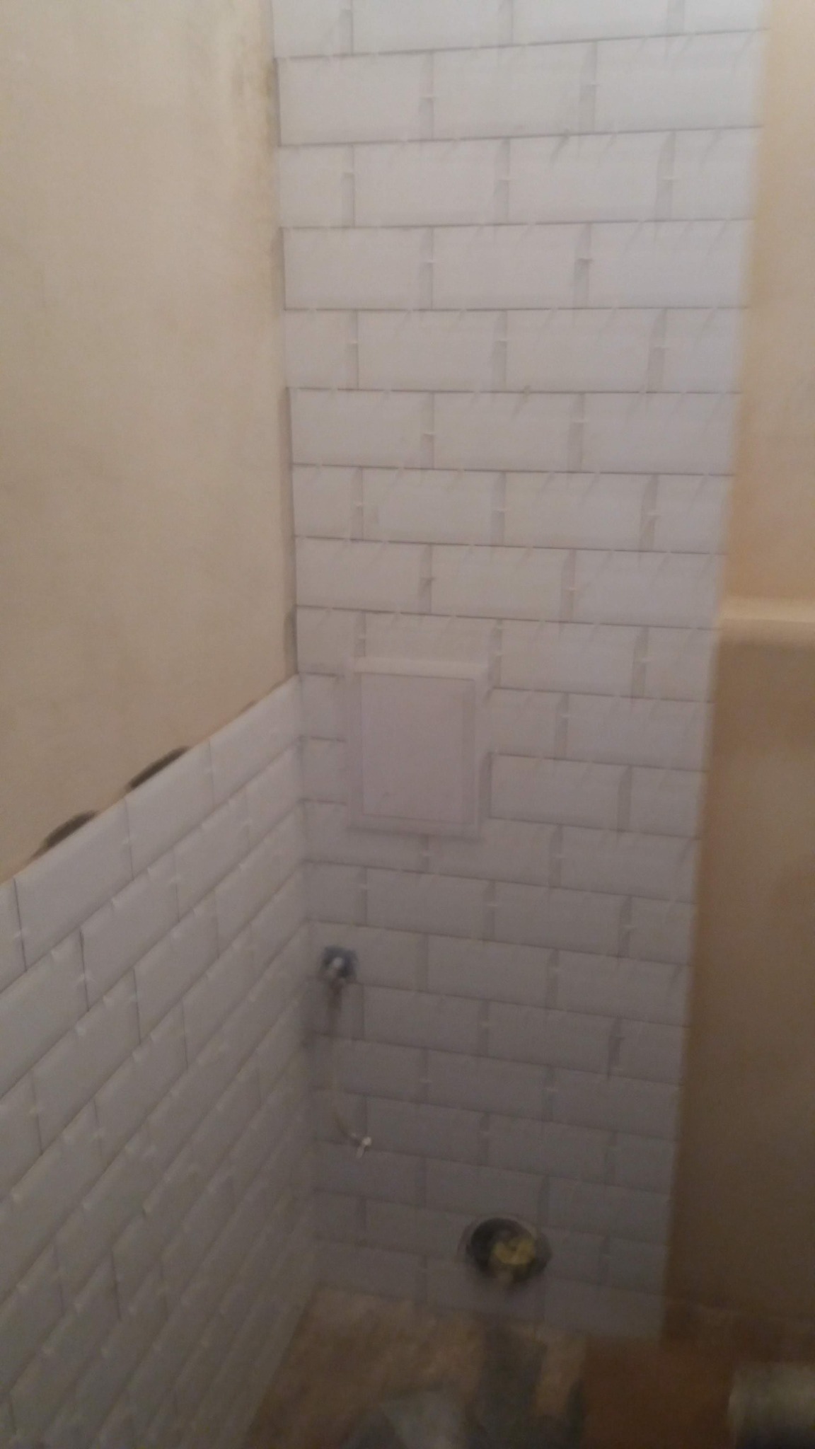 Świeżo ułożone białe, prostokątne płytki typu metro na ścianach w narożniku łazienki, z widocznym otworem rewizyjnym i surowymi ścianami w tle, ukazujące etap prac wykończeniowych.