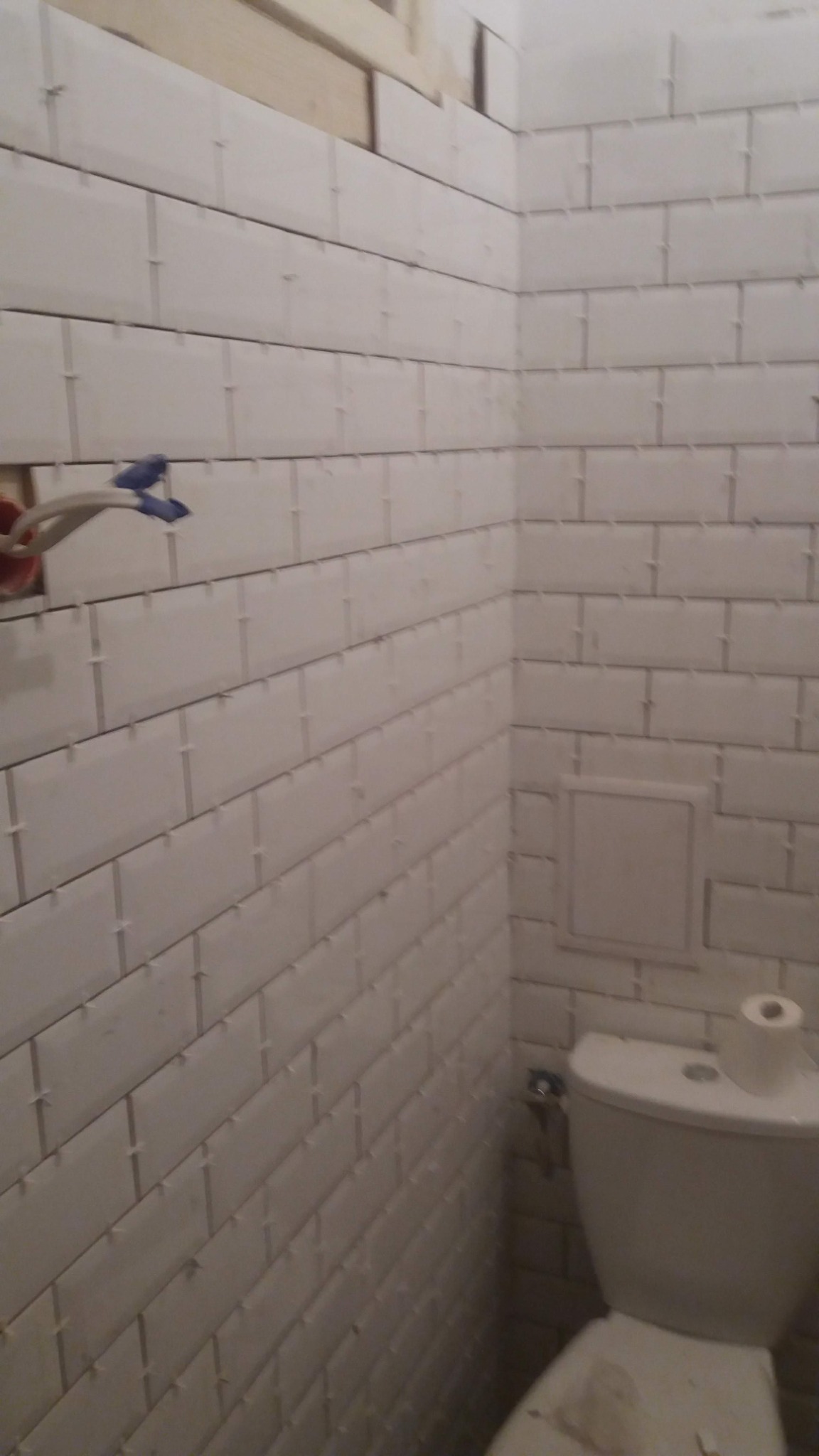 Białe, prostokątne płytki ułożone na ścianie łazienki z krzyżykami dystansowymi, widoczne fragmenty instalacji elektrycznej i częściowo zamontowana toaleta