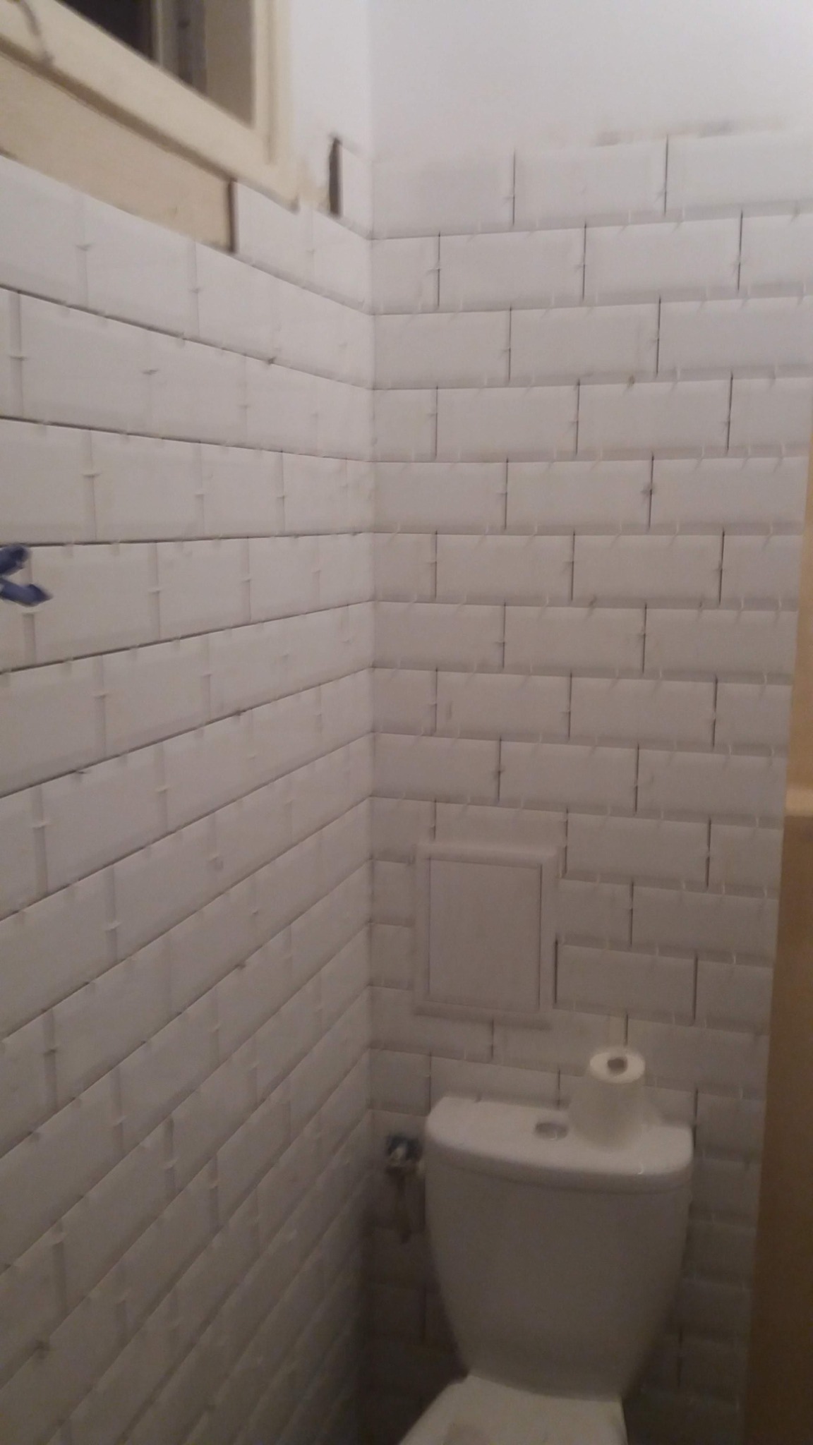 Świeżo wykafelkowana ściana łazienki z białymi, prostokątnymi płytkami ułożonymi w cegiełkę, widoczna toaleta z rolką papieru toaletowego na górze, szare fugi, nieukończone wykończenie okna.