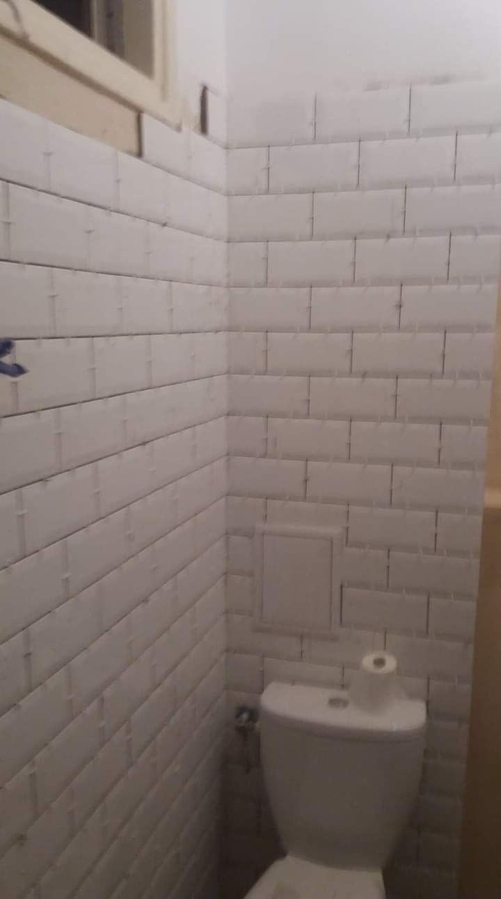 Ściana łazienki wyłożona białymi, prostokątnymi płytkami, z widocznym oknem u góry i toaletą z rolką papieru, ukazująca ukończone prace.