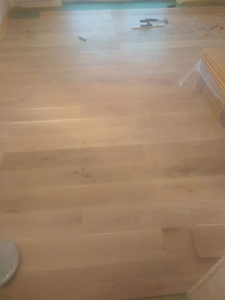 Jasne, drewniane panele podłogowe w trakcie montażu, widoczne narzędzia i paczka paneli w rogu pomieszczenia.