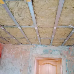 Poddasze w trakcie ocieplania wełną mineralną, widoczne metalowe profile sufitowe i przewody elektryczne zwisające ze stropu, ściany w stanie surowym z widocznymi śladami farby.