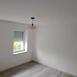 Pusty, jasny pokój po remoncie z dwoma oknami z roletami, drewnopodobną podłogą i nowoczesną, bursztynową lampą wiszącą.