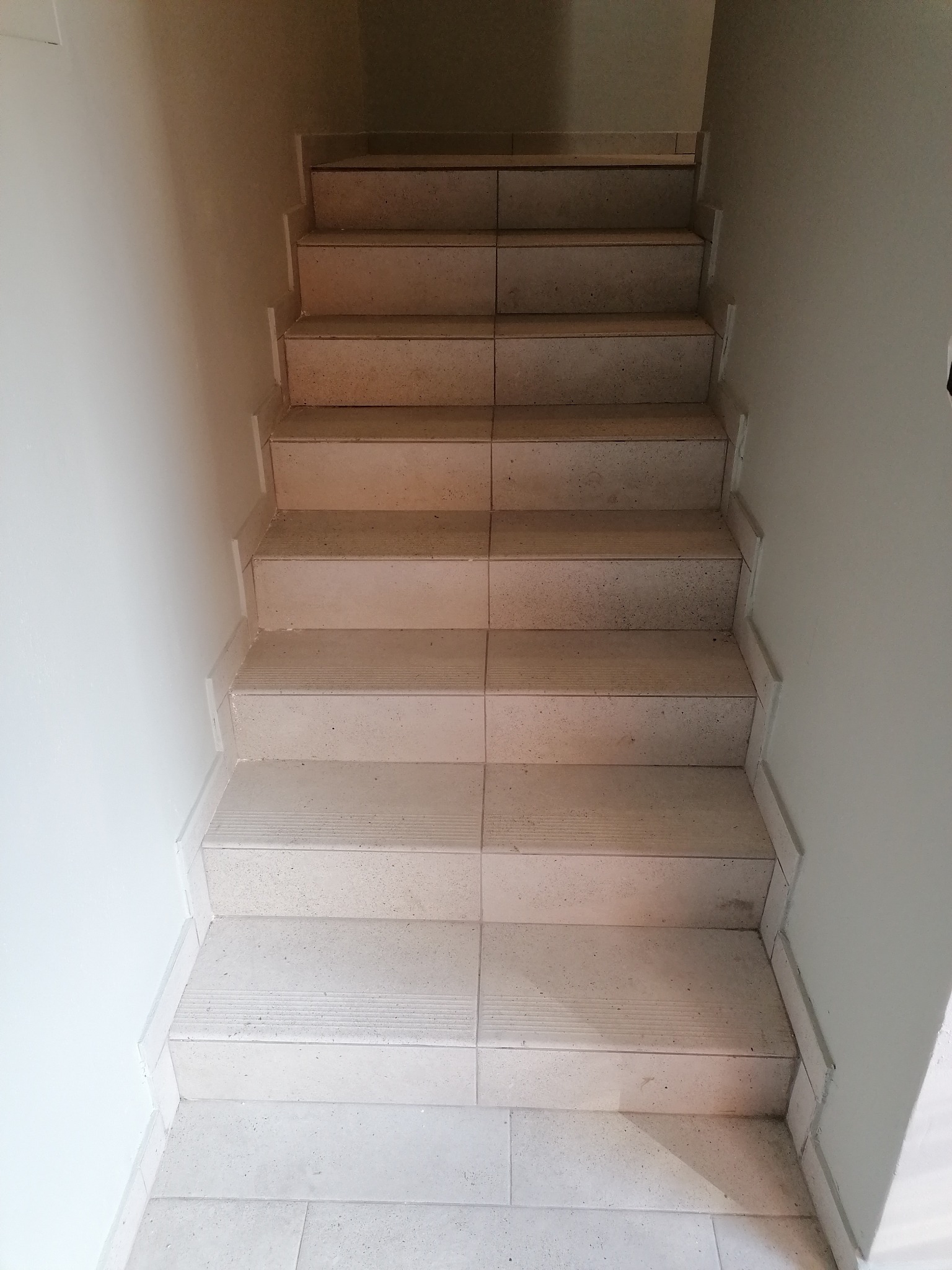 Schody wyłożone jasnymi, kwadratowymi płytkami ceramicznymi, widok z góry na dół, ściana pomalowana na jasny kolor.