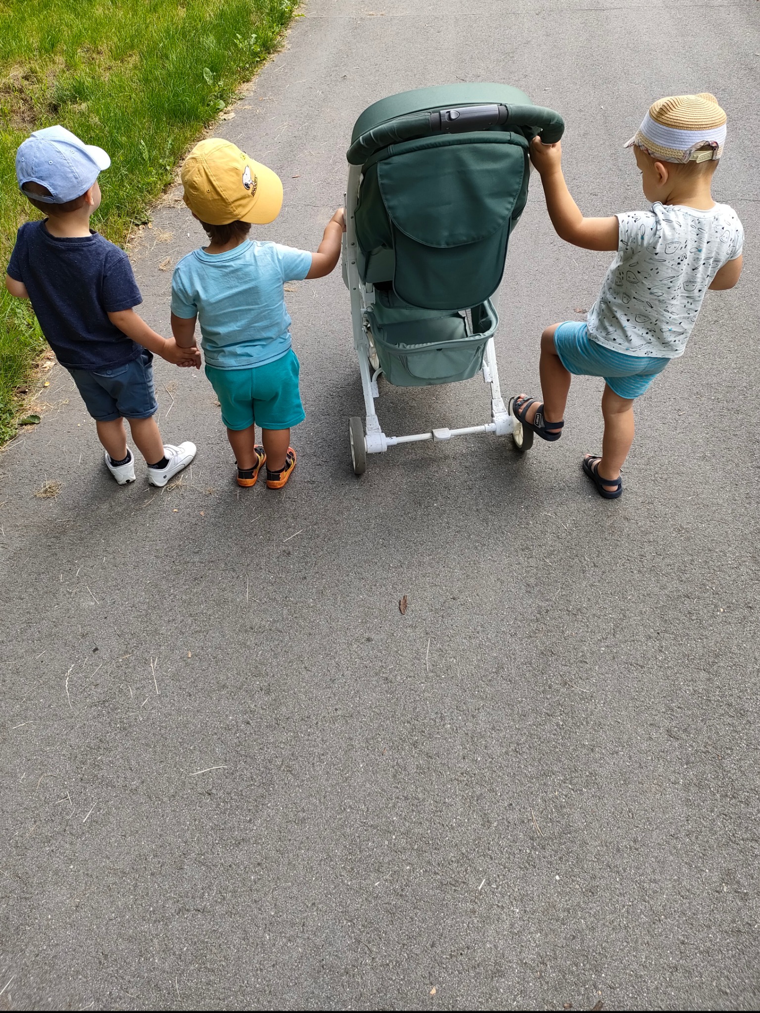 Trójka dzieci w kapeluszach, trzymająca się za ręce i pchająca zielony wózek dziecięcy na chodniku.