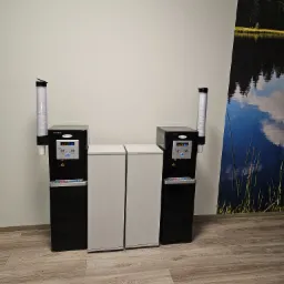 Dwa dystrybutory wody marki Ruhens z pełnymi zasobnikami na kubki, umieszczone obok dwóch białych szafek w biurze, z fototapetą przedstawiającą jezioro i las w tle.