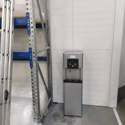 P.O.U. AQUA WARMIA Marta Magdaleńska - Srebrny dystrybutor wody z podajnikiem kubków, ustawiony w magazynie w Olsztynie, obok regałów z towarem i drabiny.