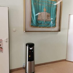 P.O.U. AQUA WARMIA Marta Magdaleńska - Srebrny dystrybutor wody z czarnym panelem, umieszczony w korytarzu szkolnym pod oprawionym sztandarem.