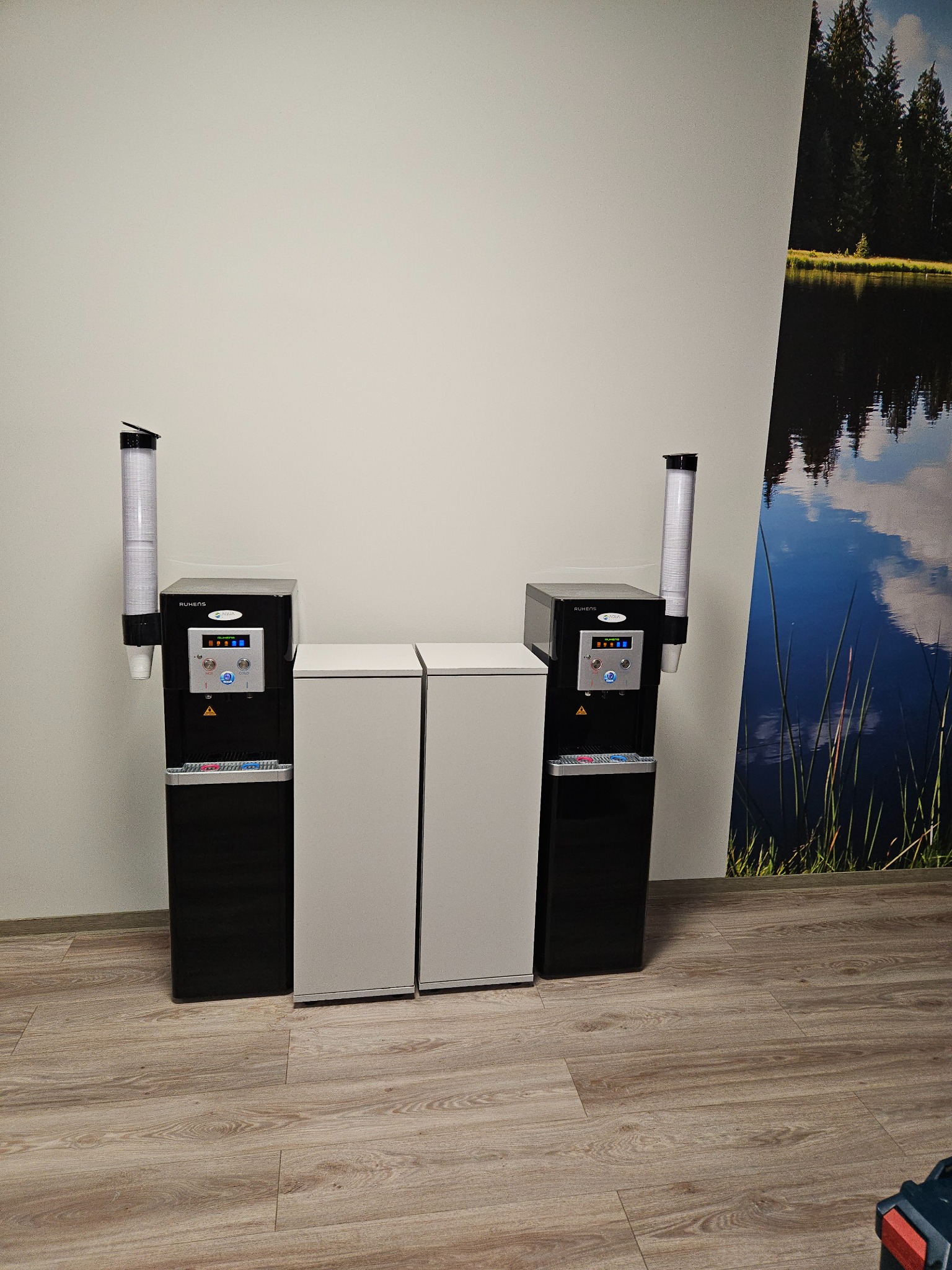 Dwa dystrybutory wody marki Ruhens z pełnymi zasobnikami na kubki, umieszczone obok dwóch białych szafek w biurze, z fototapetą przedstawiającą jezioro i las w tle.