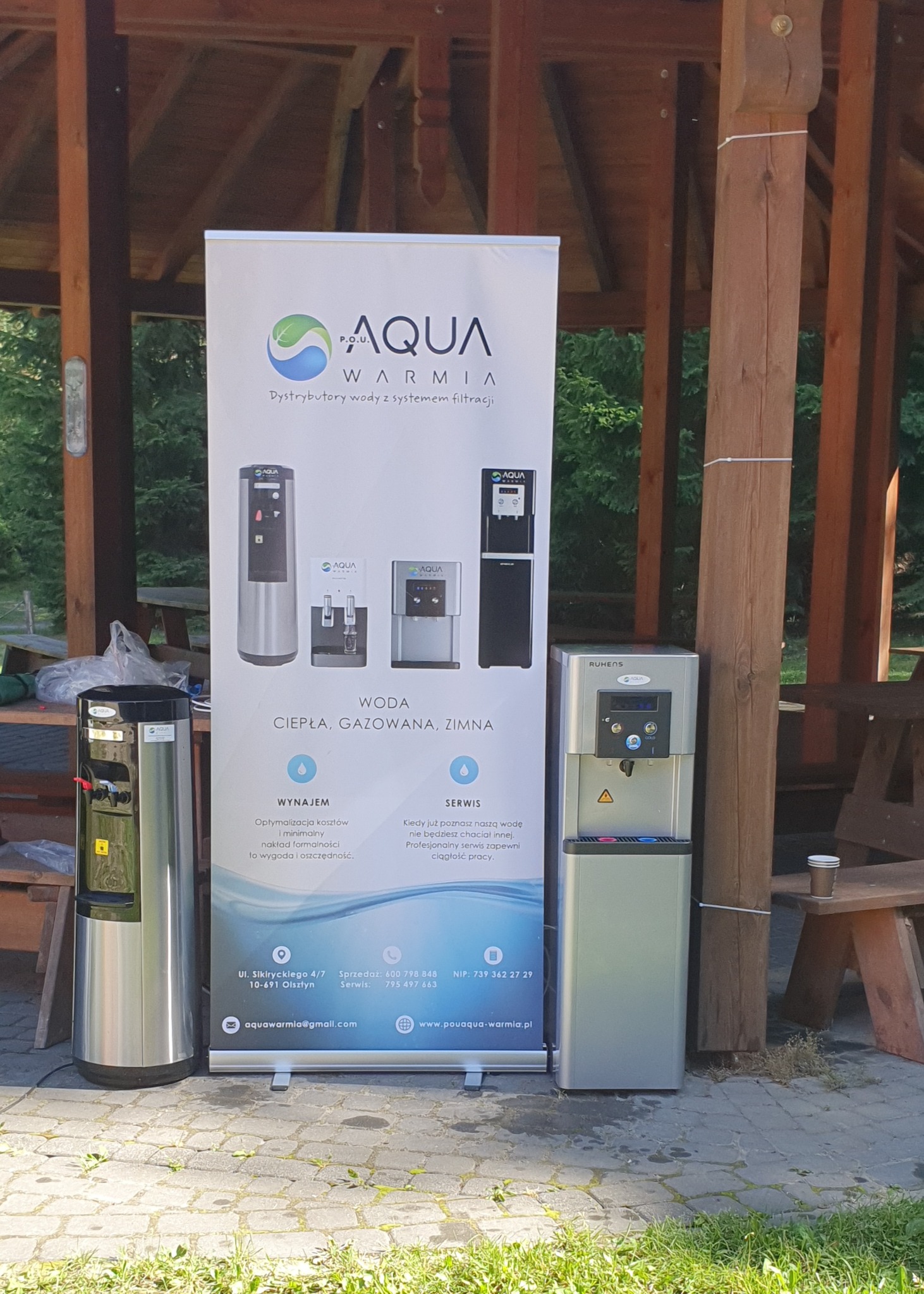 Prezentacja dystrybutorów wody Aqua Warmia, z trzema modelami urządzeń obok baneru reklamowego, na zewnątrz w zadaszonej altanie.