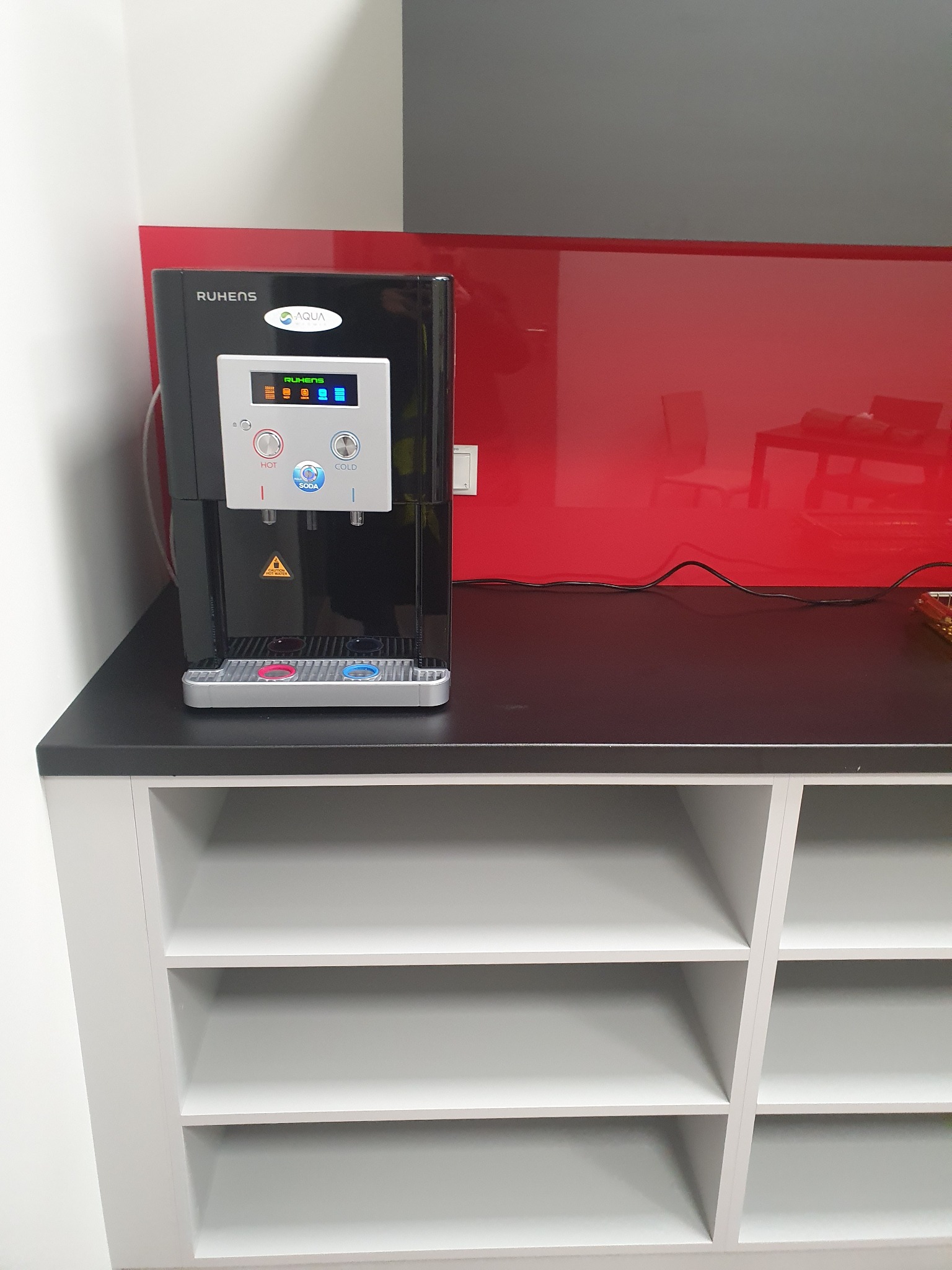 Czarny dystrybutor wody Ruhens Aqua na blacie z szarą szafką, czerwone szkło na ścianie w tle.
