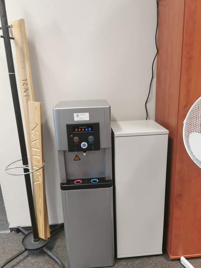 Srebrny dystrybutor wody Aqua stojący obok białej szafki i wieszaka na ubrania z drewnianymi elementami dekoracyjnymi w biurze.