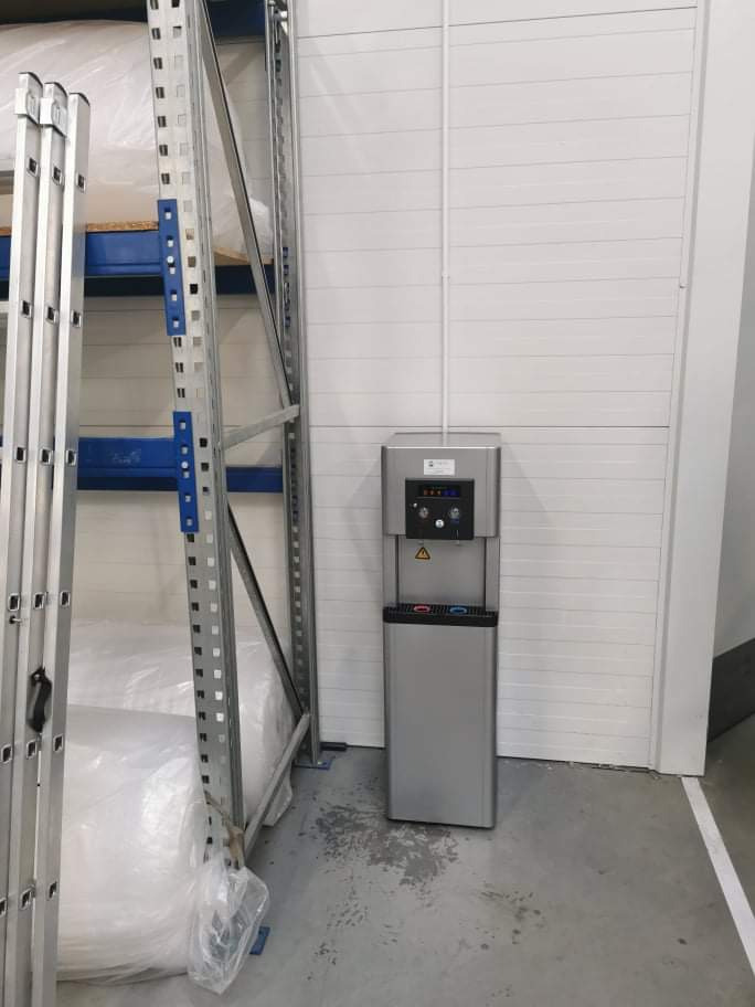 Srebrny dystrybutor wody z podajnikiem kubków, ustawiony w magazynie w Olsztynie, obok regałów z towarem i drabiny.