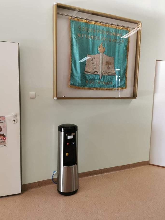 Srebrny dystrybutor wody z czarnym panelem, umieszczony w korytarzu szkolnym pod oprawionym sztandarem.