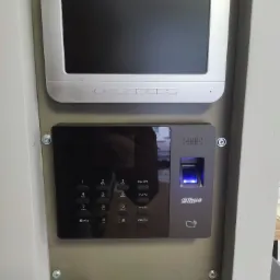 Zamontowany wideodomofon Hikvision i czytnik linii papilarnych Dahua z podświetlonym skanerem, przymocowane do ściany za pomocą śrub, z kartką 'NILON SOPST' w dolnej części.