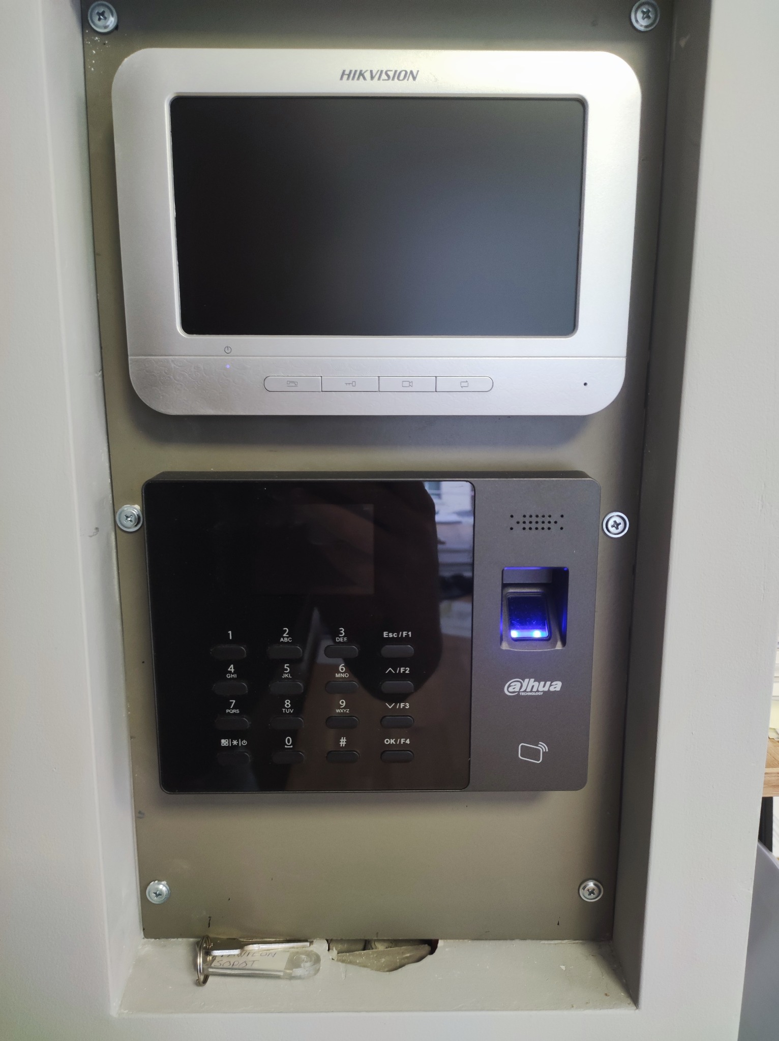 Zamontowany wideodomofon Hikvision i czytnik linii papilarnych Dahua z podświetlonym skanerem, przymocowane do ściany za pomocą śrub, z kartką 'NILON SOPST' w dolnej części.