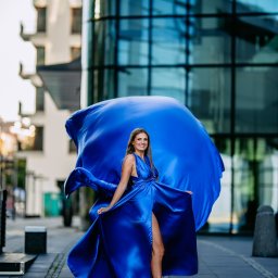 Art Foto Katarzyna Margol - Kobieta w długiej, powiewnej, kobaltowej sukni pozuje na tle nowoczesnego budynku z przeszkleniami, uśmiechając się do kamery.