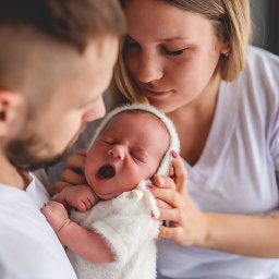Art Foto Katarzyna Margol - Rodzice tulą noworodka w białym stroju, dziecko ziewa. Bliski kadr, miękkie światło, rodzinna sesja w studio. Widoczne dłonie rodziców i małe rączki dziecka.