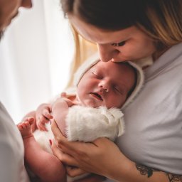 Art Foto Katarzyna Margol - Mama całuje śpiące niemowlę ubrane w biały, futerkowy kombinezon, w ramionach rodziców. Delikatne światło, widoczne tatuaże na ramieniu matki.
