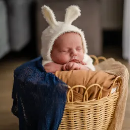 Śpiący noworodek w kostiumie królika leży w wiklinowym koszu, otulony niebieską i beżową tkaniną. Delikatne, naturalne światło podkreśla słodki wyraz twarzy dziecka.