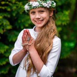 Art Foto Katarzyna Margol - Uśmiechnięta dziewczynka w białej sukience komunijnej, z wiankiem na głowie, trzymająca różaniec na tle zieleni. Portret w naturalnym świetle, z widocznym logo fotografa.