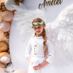 Art Foto Katarzyna Margol - Uśmiechnięta dziewczynka w białej sukience komunijnej z wiankiem, pozuje na tle dekoracji z balonów i skrzydeł anielskich. W rękach trzyma świecę.