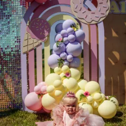 Roczne dziecko w różowej sukience siedzi na kocu na trawie, z dekoracjami urodzinowymi w tle: balony, tęcza, donut i lody. Ujęcie z reportażu urodzinowego.