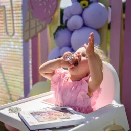 Radosna dziewczynka w różowej sukience pije z małej szklanki na krzesełku, otoczona dekoracjami urodzinowymi w odcieniach fioletu i różu, w plenerze.