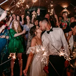 Pocałunek nowożeńców otoczonych gośćmi z zimnymi ogniami na tle ceglanej ściany z neonowym napisem 'Progres'.