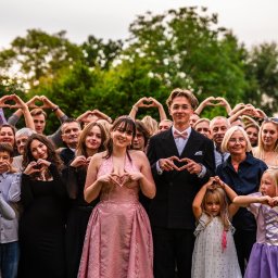 Art Foto Katarzyna Margol - Grupowe zdjęcie z uroczystości, goście tworzą serca dłońmi. W centrum para w strojach wieczorowych, dziewczyna w różowej sukni, chłopak w garniturze z muszką.