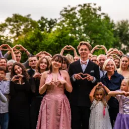 Grupowe zdjęcie z uroczystości, goście tworzą serca dłońmi. W centrum para w strojach wieczorowych, dziewczyna w różowej sukni, chłopak w garniturze z muszką.