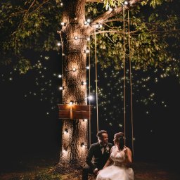 Art Foto Katarzyna Margol - Para młoda siedzi na huśtawce pod rozświetlonym drzewem w nocy. Romantyczna sesja ślubna w plenerze, ciepłe światło lampek.