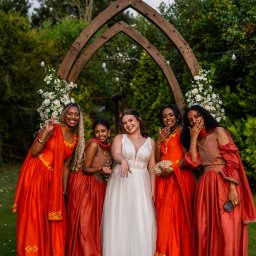 Art Foto Katarzyna Margol - Panna młoda w białej sukni i druhny w pomarańczowych sukniach pozują pod drewnianym łukiem udekorowanym kwiatami. Zdjęcie wykonane w Gdyni.