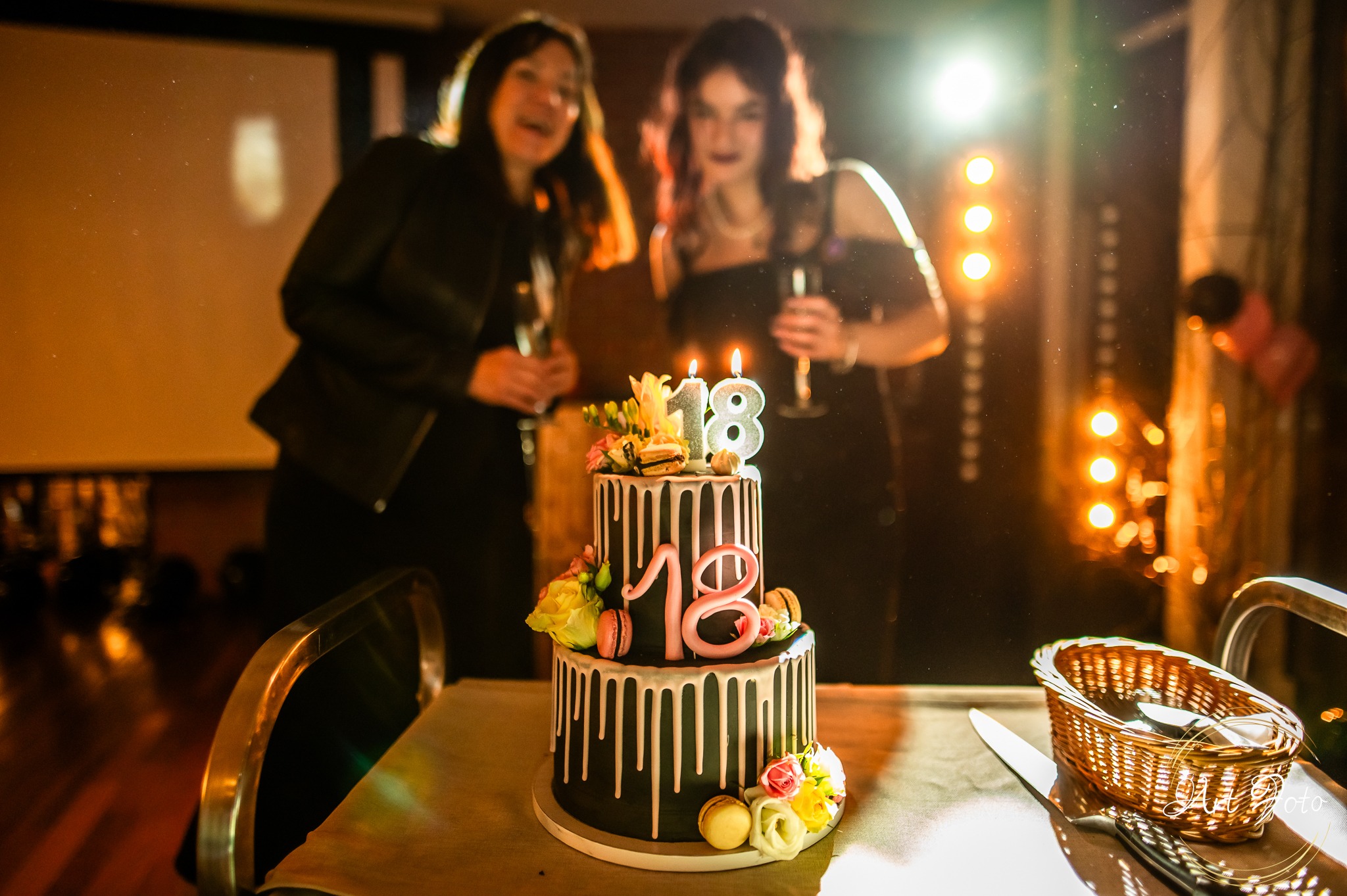 Uroczysty tort osiemnastkowy z płonącymi świeczkami na stole; w tle dwie kobiety z kieliszkami w dłoniach, oświetlenie sceniczne. Dekoracje kwiatowe i makaroniki.