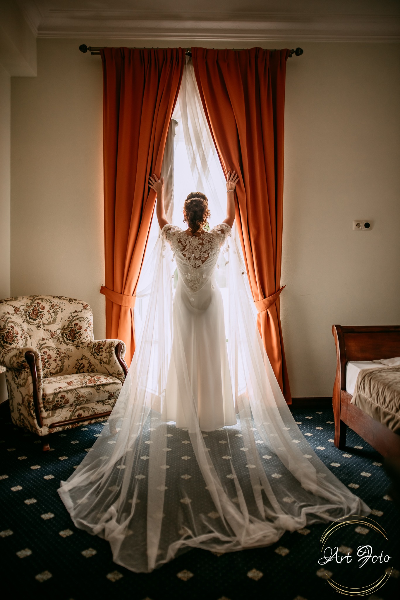 Panna młoda w koronkowej sukni i długim welonie stoi tyłem przy oknie z pomarańczowymi zasłonami w pokoju z wzorzystym fotelem i łóżkiem.