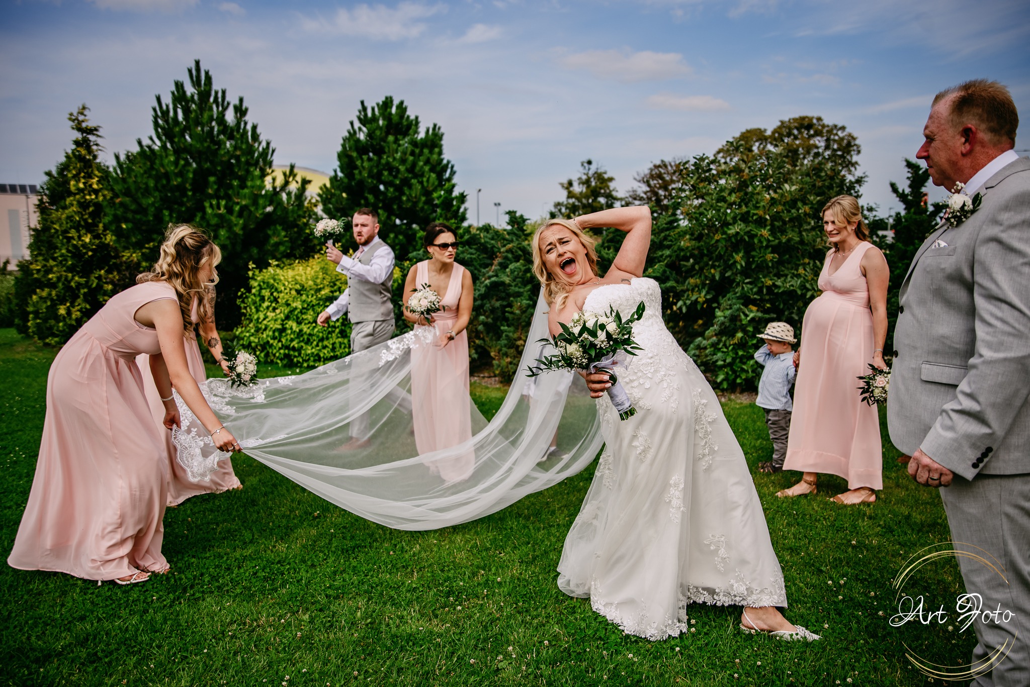 Rozbawiona panna młoda w koronkowej sukni pozuje z druhnami w różowych sukienkach i gośćmi na zielonym trawniku podczas sesji ślubnej w plenerze.