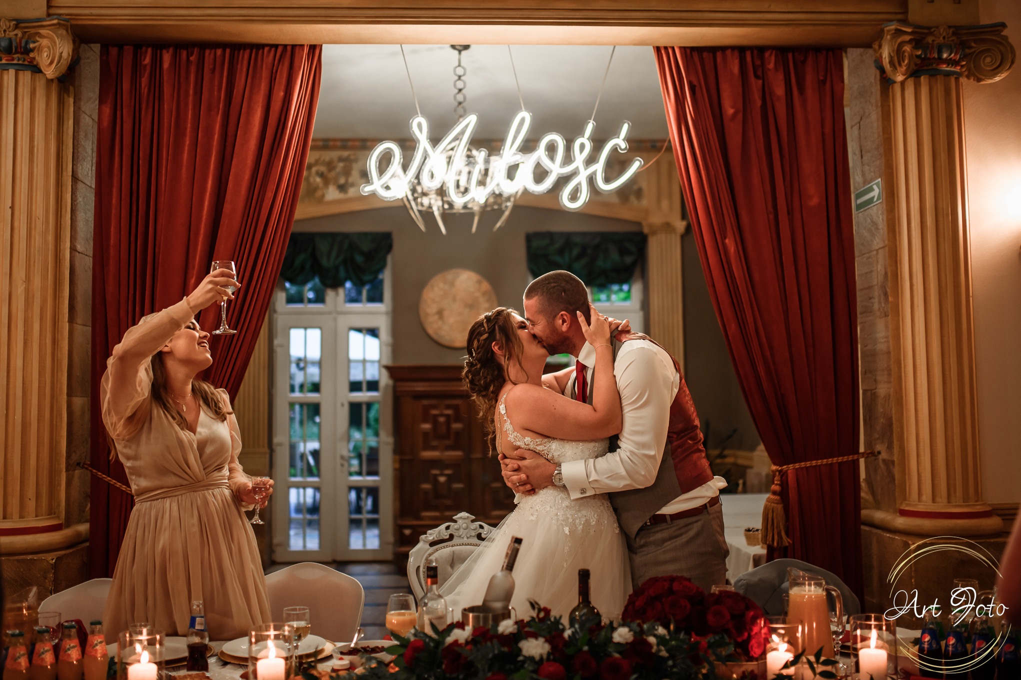 Pocałunek pary młodej pod neonem 'Miłość' na sali weselnej z czerwoną dekoracją. Gość wznosi toast. Ujęcie z dołu, ciepłe światło.