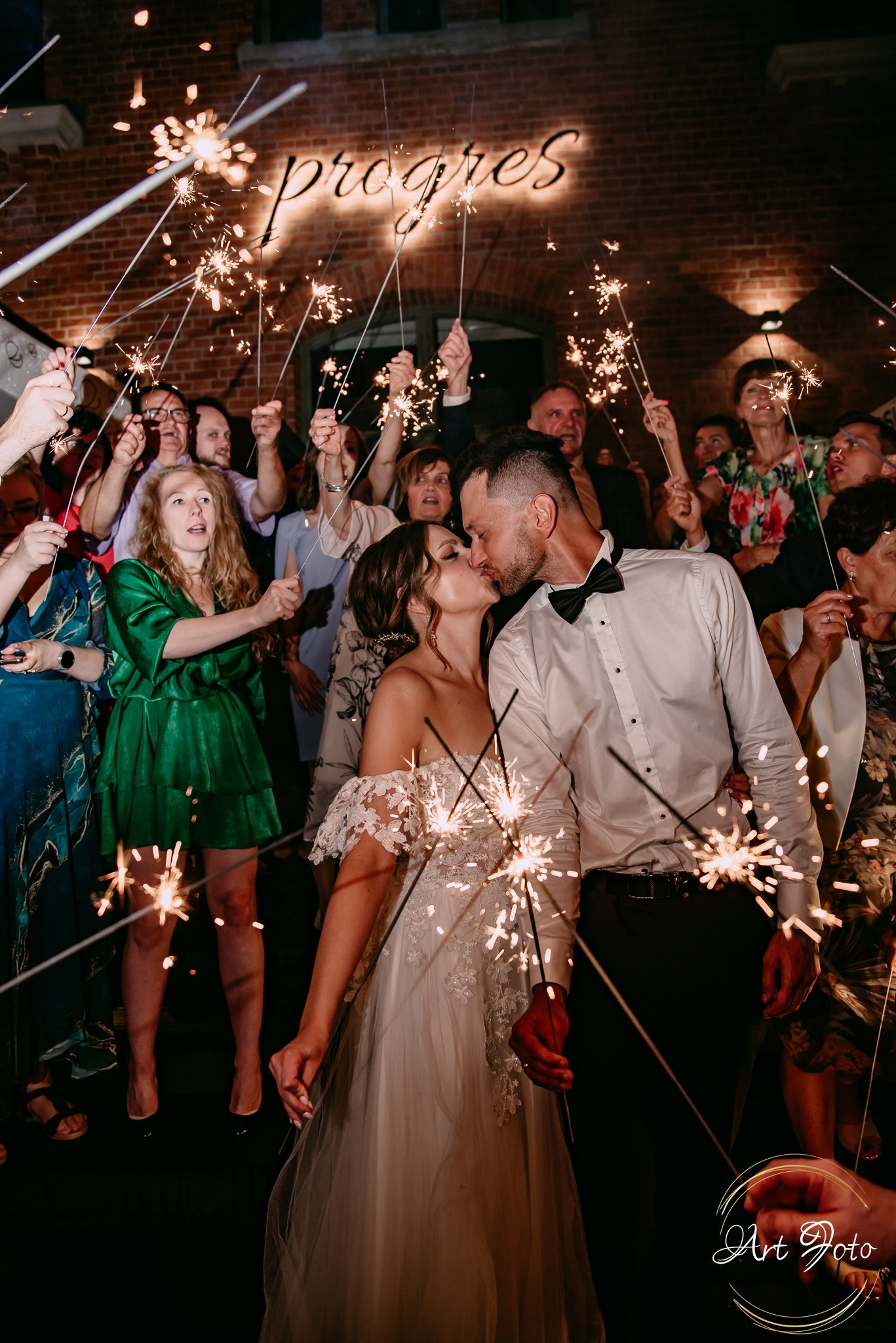 Pocałunek nowożeńców otoczonych gośćmi z zimnymi ogniami na tle ceglanej ściany z neonowym napisem 'Progres'.