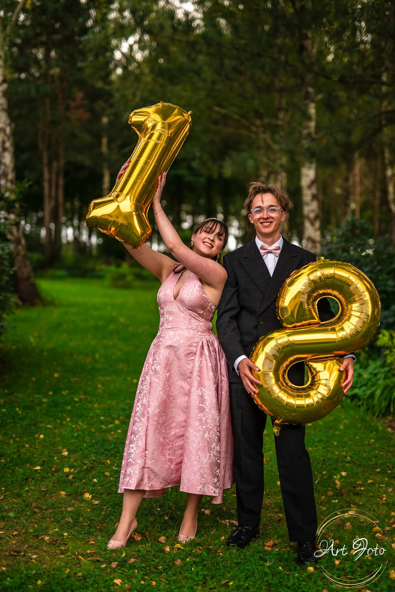 Uśmiechnięta para trzyma złote balony w kształcie cyfr 1 i 8 na tle leśnej zieleni. Dziewczyna w różowej sukni, chłopak w garniturze z muszką. Okazja: 18 urodziny.