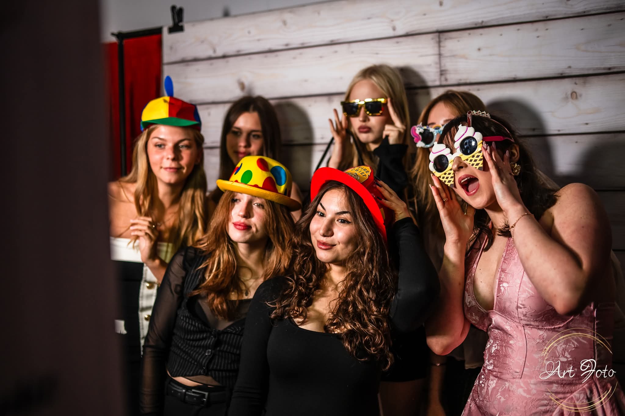 Grupa kobiet w zabawnych przebraniach pozuje do zdjęcia w fotobudce. Kolorowe kapelusze, okulary i radosne miny tworzą wesołą atmosferę imprezy.