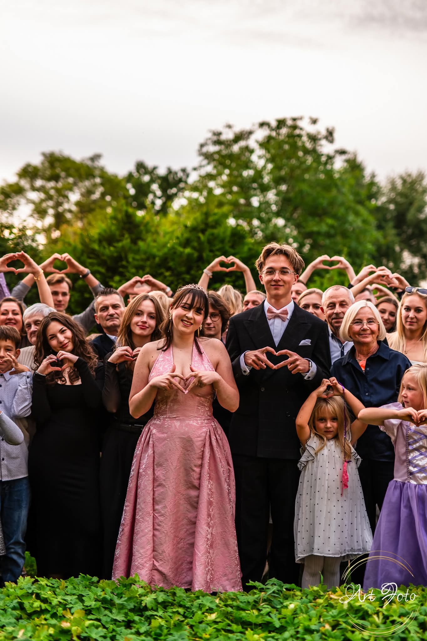 Grupowe zdjęcie z uroczystości, goście tworzą serca dłońmi. W centrum para w strojach wieczorowych, dziewczyna w różowej sukni, chłopak w garniturze z muszką.