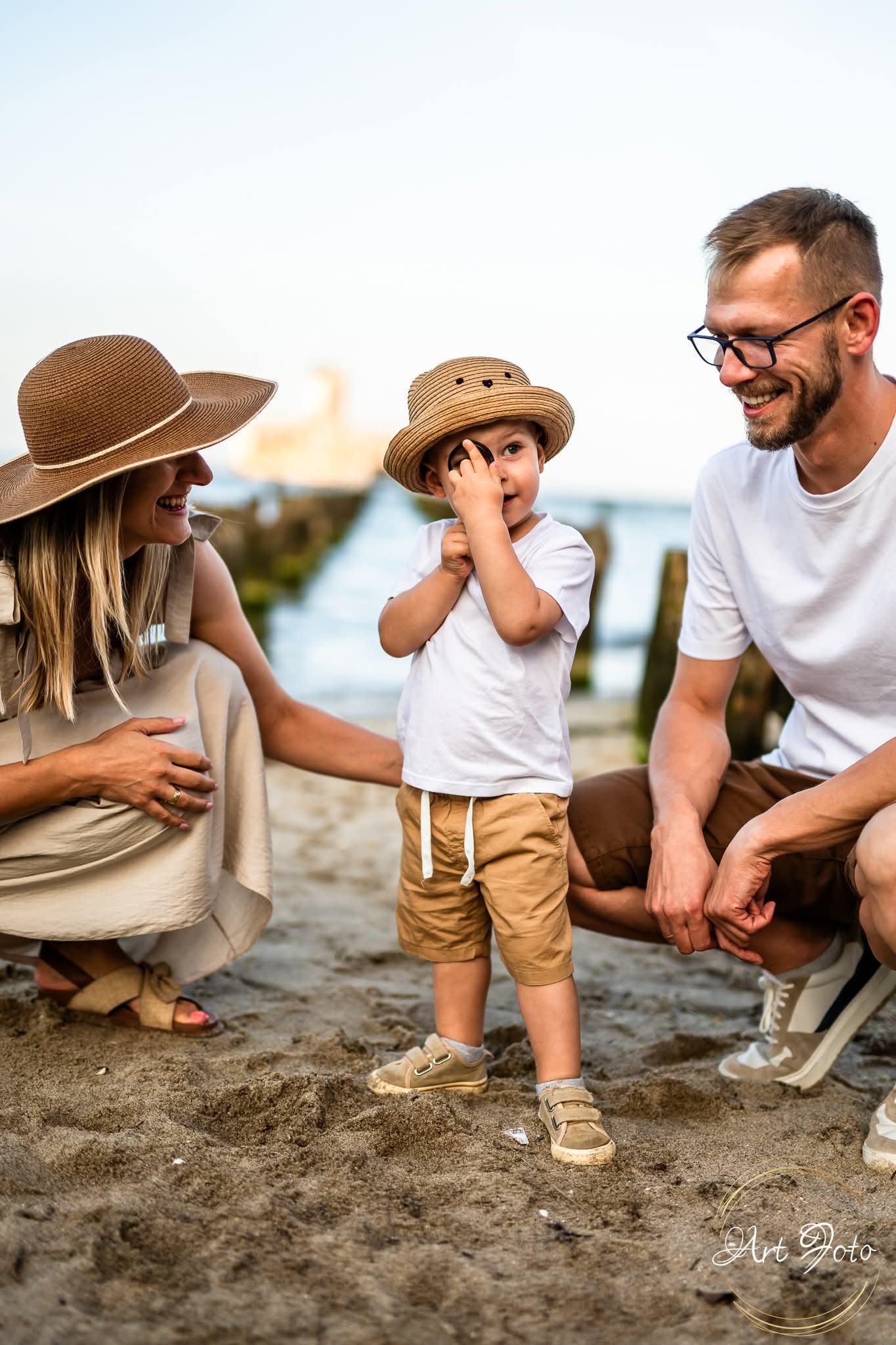 Uśmiechnięta rodzina na plaży: mama w kapeluszu, tata w okularach i syn w kapeluszu z dziurkami, bawiący się dłońmi przed okiem. W tle woda i drewniane pale.