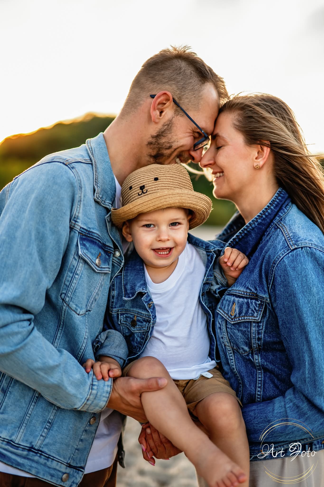 Uśmiechnięta rodzina: tata w okularach, mama i synek w kapeluszu z uszami misia, w jeansowych kurtkach, tulą się do siebie na tle zachodzącego słońca.