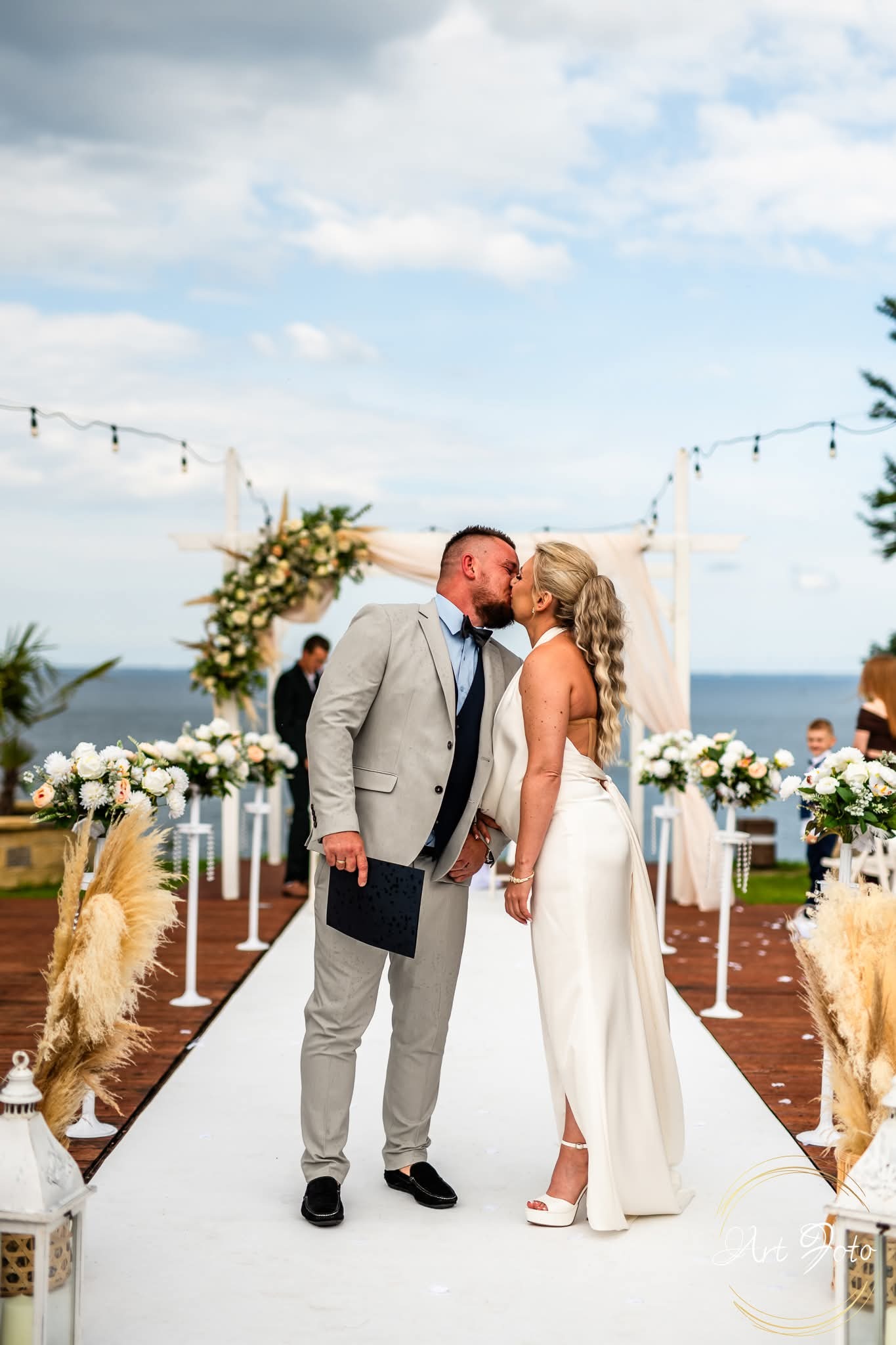 Pocałunek pary młodej na tle dekoracji ślubnej z widokiem na morze. Pan młody w szarym garniturze trzyma czarny dokument. Panna młoda w białej sukni z odkrytymi plecami.