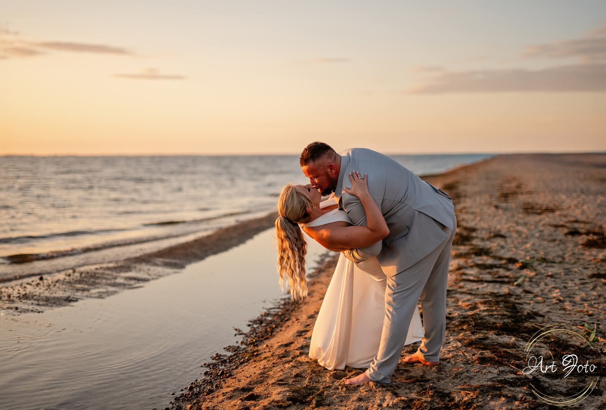 Para młoda w objęciach na plaży o zachodzie słońca. Panna młoda w białej sukni, pan młody w szarym garniturze. Romantyczny pocałunek na tle morza i nieba w ciepłych barwach.