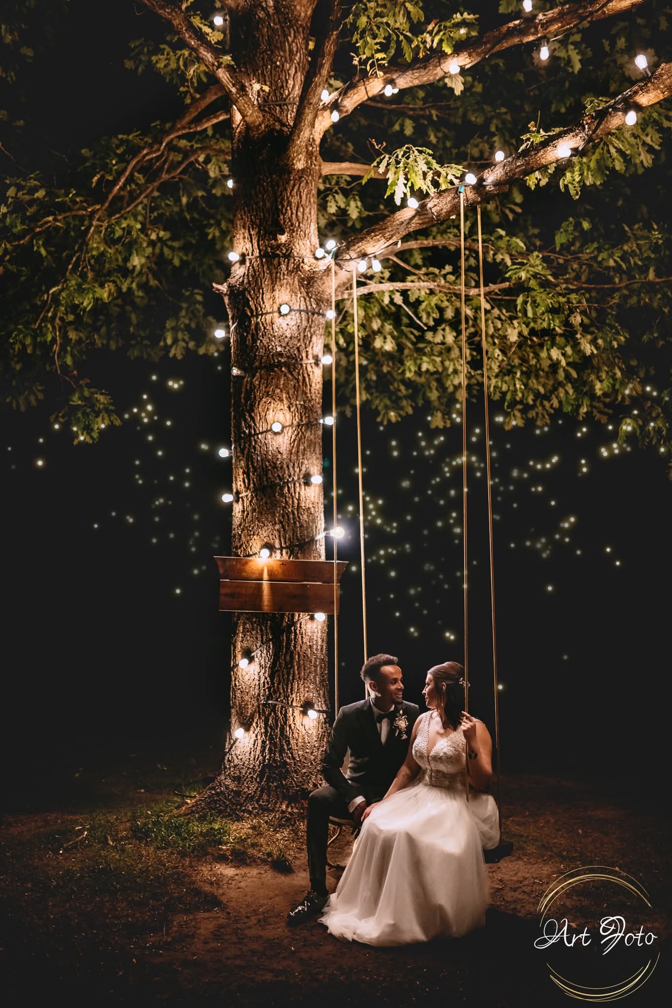 Para młoda siedzi na huśtawce pod rozświetlonym drzewem w nocy. Romantyczna sesja ślubna w plenerze, ciepłe światło lampek.