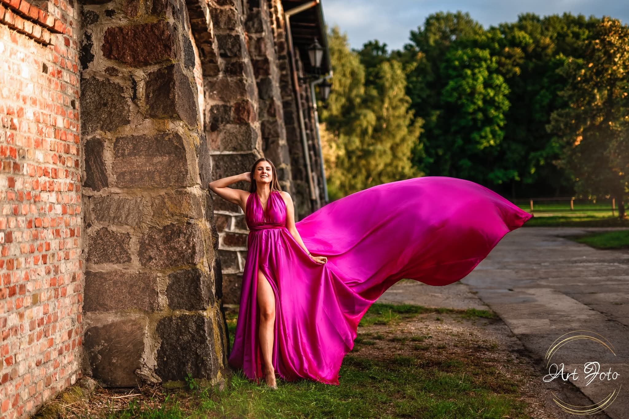 Uśmiechnięta kobieta w fuksjowej sukni z rozcięciem, oparta o kamienny mur. Suknia powiewa na wietrze, tło to zieleń drzew i ścieżka.